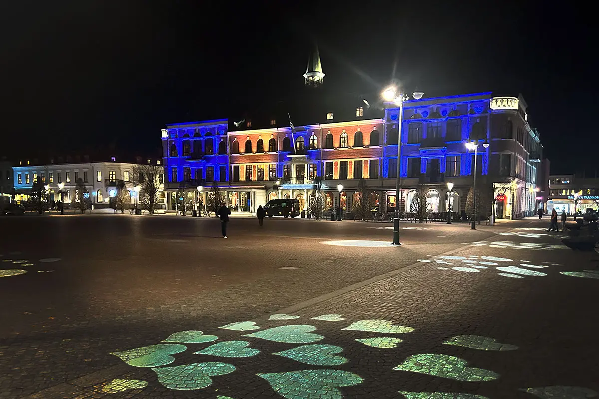Varbergs Stadshotell i kvällsbelysning. Fasaden har en belysning i blått och gult. Torget syns i förgrunden och kullerstenen har belysta hjärtan på marken.