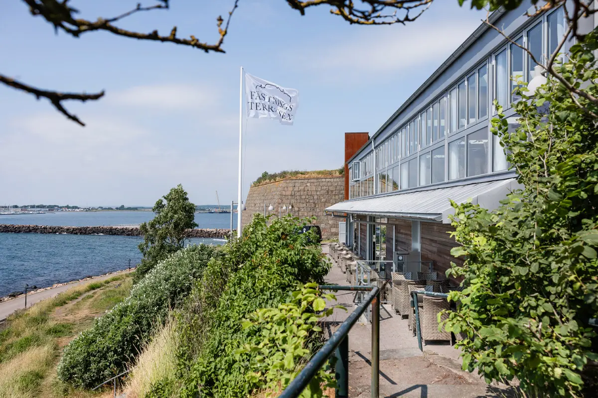 Bild tagen över uteserveringen som löper längs hela byggnadens långsida. I bakgrunden en vit flagga med logo, strandpromenaden och havet.