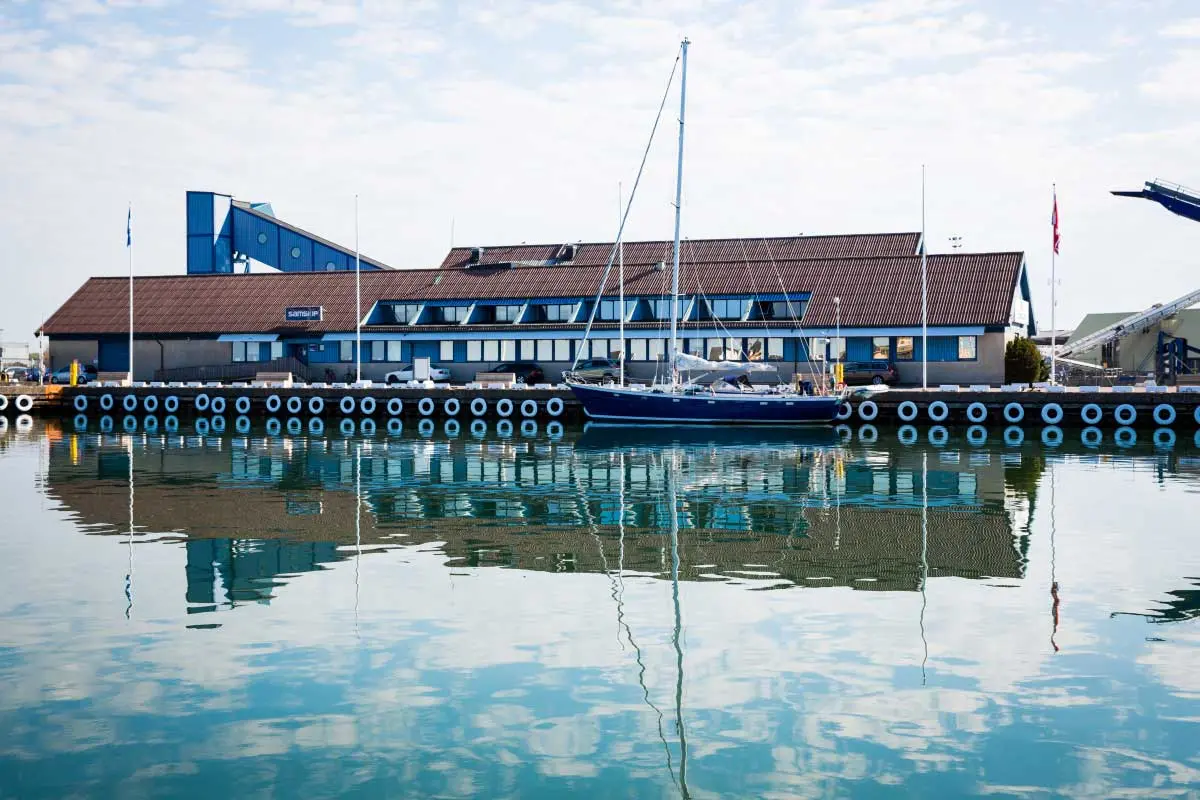 Den gamla färjeterminalen i Varbergs hamn. Långsträckt byggnad med många fönster speglas i hamnbassängens vatten. En segelbåt ligger förtöjd.