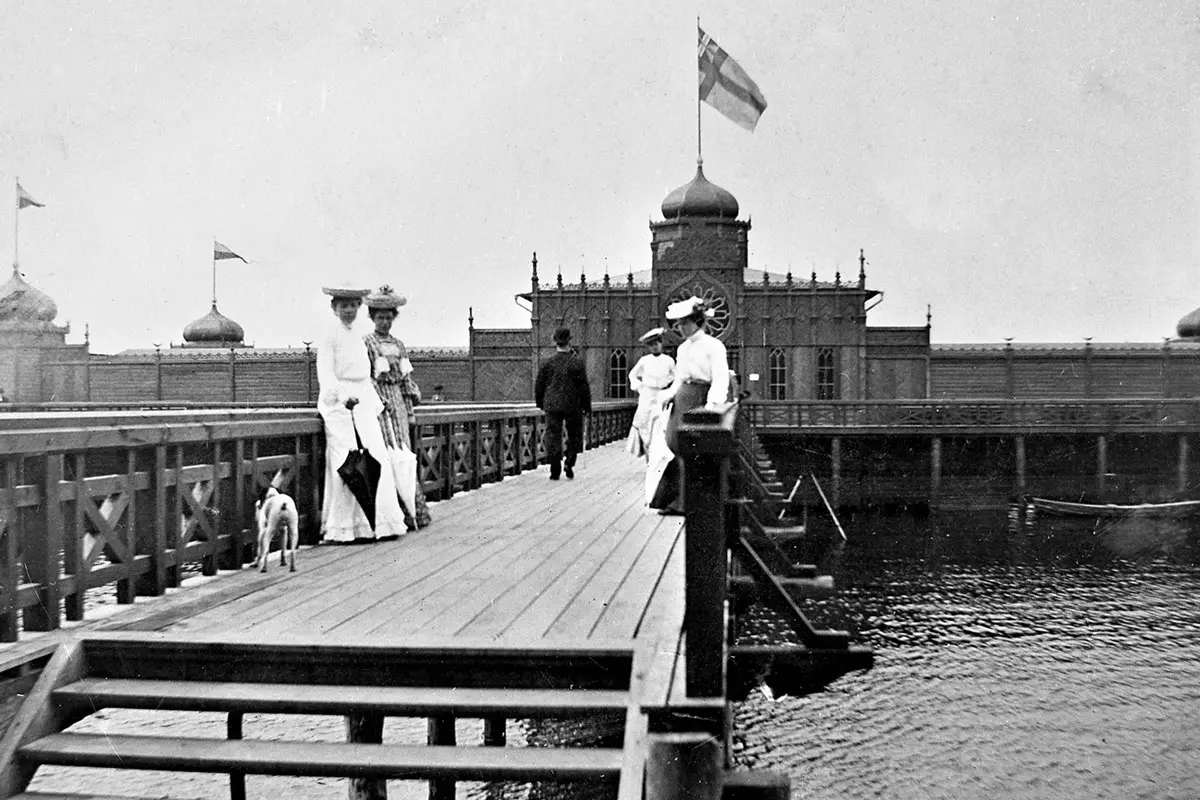 Svartvitt fotot. Badgäster med vita kläder och hattbeklädnad står på bryggan ut till Kallbadhuset. 