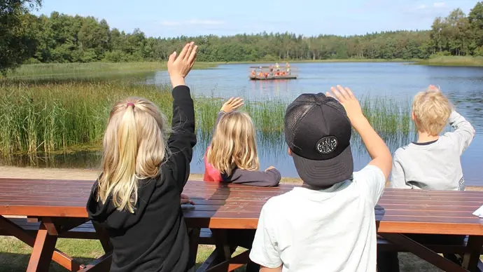 I förgrunden barn runt träbord. Barnen vinkar till personer ute på flotten i sjön.