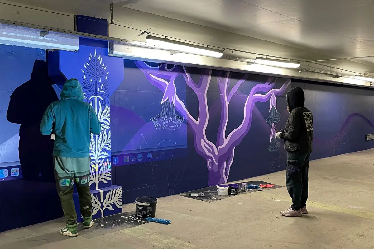Två personer målar träd och blommor på en mörkblå betongvägg.