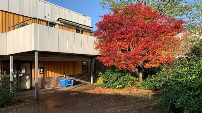 En entré med balkong ovanför. Byggnaden har mörkt gul fasad med inslag av vita plåtskivor. Ett stort träd med rödorange höstfärg på löven.
