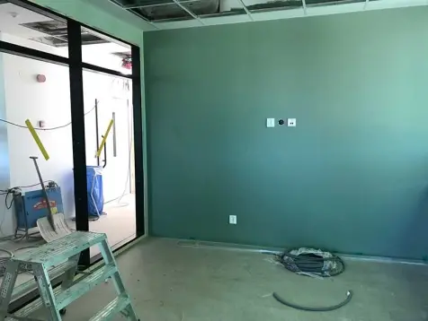 Rum med gröna väggar och grått golv. Väggen till vänster består av glaspartier. I rummet står en byggstege.