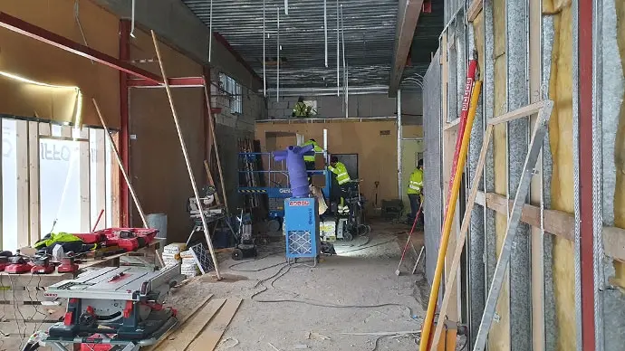 Rum fyllt av byggmaterial. En blå värmefläkt i bildens mitt. Arbetare klättrar på en stege.