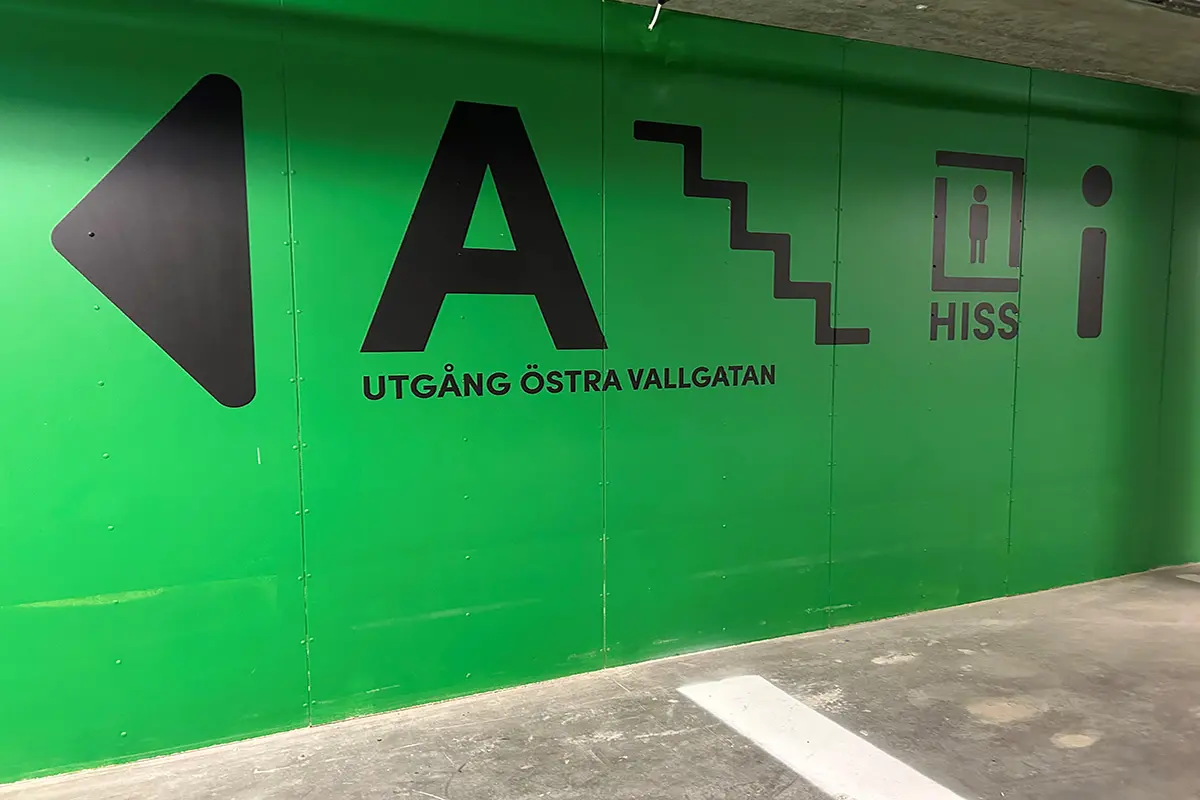 Ett parkeringsgarage med en grönmålad vägg. På väggen avbildas ikoner av pil, bokstäver, trappa och hiss.