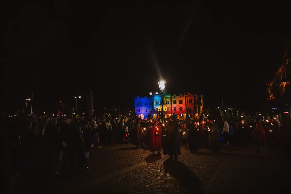 En folkmassa på Varbergs Torg i mörkret. Människor håller facklor i handen. I bakgrunden är en byggnad upplyst med kulört belysning.