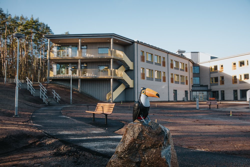 Konstverket främmande fåglar på Midsommargården. I bild, en tukan som sitter på en sten i sinnenas trädgård. I bakgrunden syns delar av en stor tegelbyggnad.