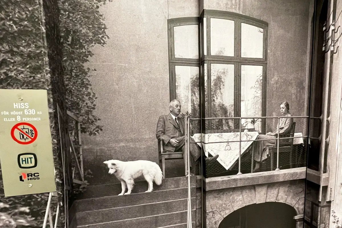 Svartvitt gammalt foto där en man och en kvinna sitter på en balkong i sten framför ett stort fönster. En vit hund står i en stentrappa som leder ner från balkongen. Till vänster i bild syns en hissknapp.
