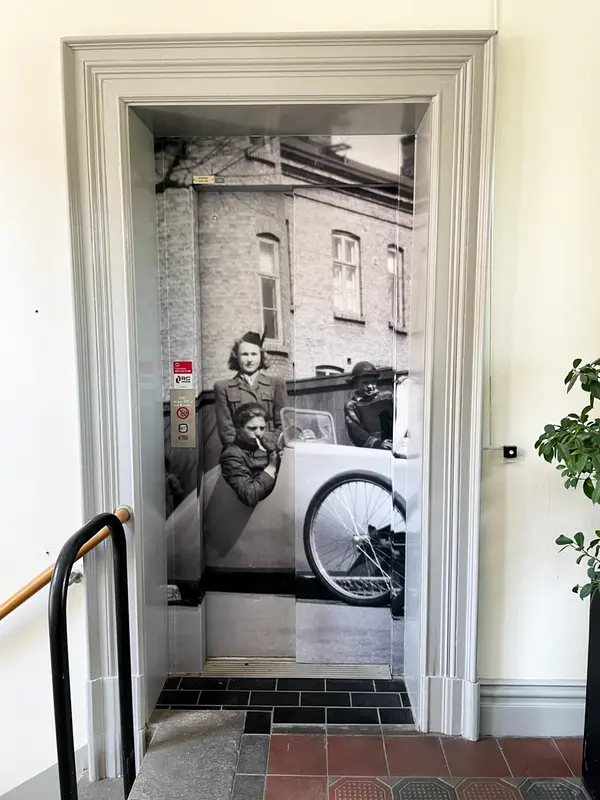En hiss där dörrarna har motiv av ett gammalt svartvitt foto där en man sitter i en lådbil med en cigarett i munnen. Bakom bilen står en kvinna i formell uniform och en pojke med skinnmössa. I bakgrunden syns ett stenhus.