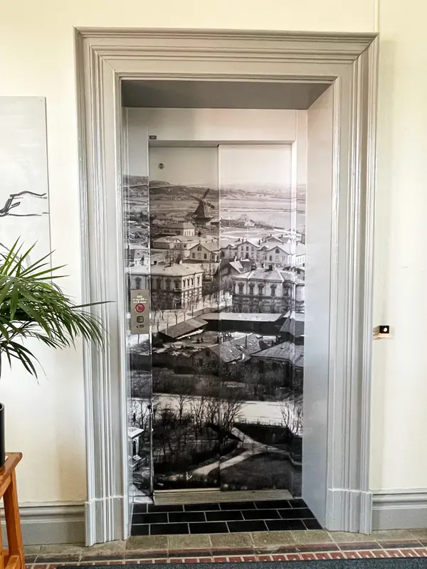 En hiss där dörrarna har motiv av ett gammalt svartvitt foto föreställandes Varberg förr i tiden. I förgrunden syns en park. Bakom den ett äldre kvarter med låghus och tvillinghusen i sten. I bakgrunden syns en stor kvarn och landskap. 