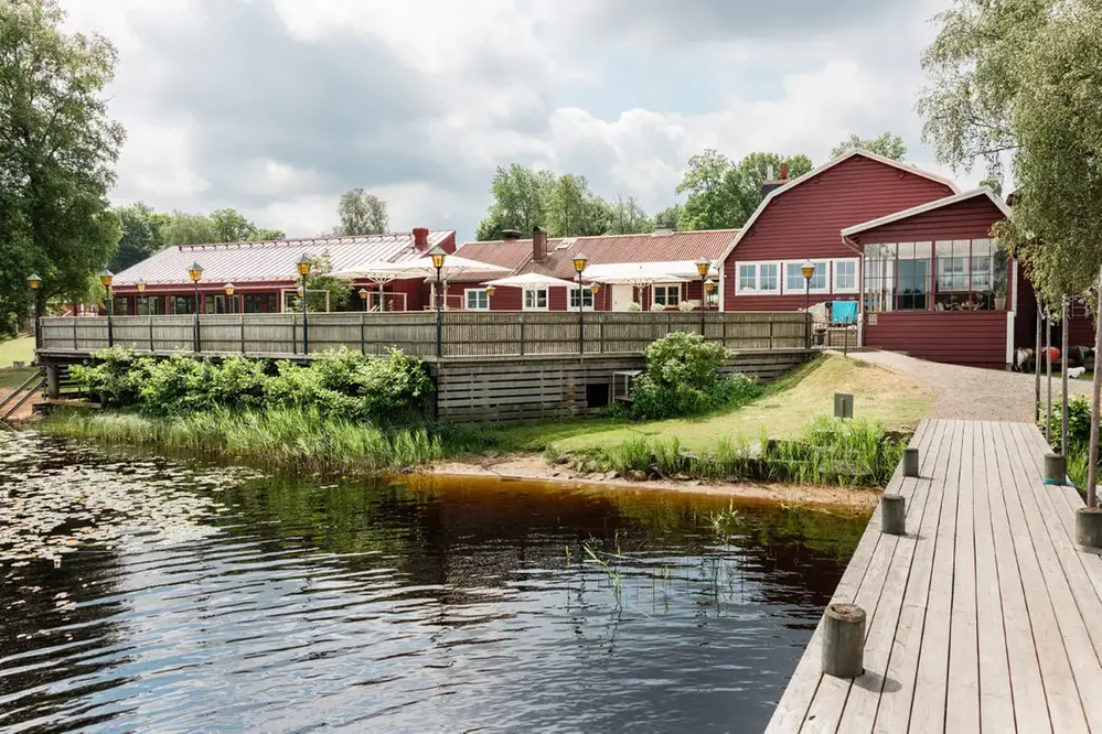 Friluftsgården och hotelldelen från bryggan. Röd byggnad i gammalsvensk stil med stor träterrass.